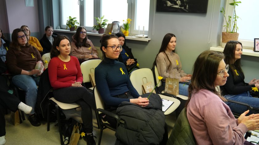 Uczestniczki spotkania siedzą na krzesłach i słuchają prelekcji, wiele z nich ma przypięte żółte wstążki i trzyma żonkile