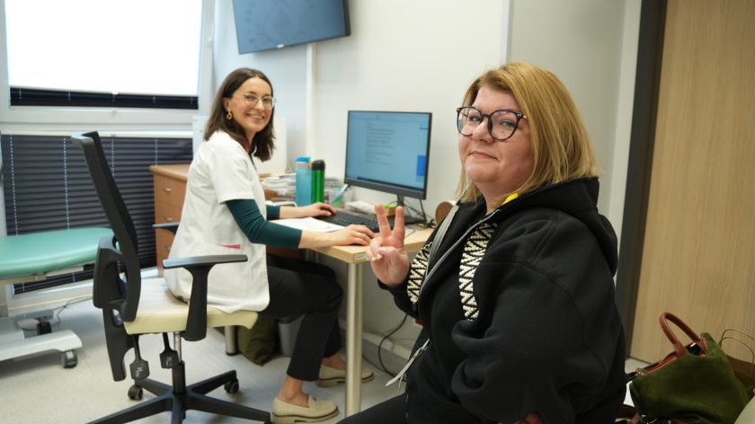 Pacjentka siedzi w gabinecie lekarskim i pokazuje znak zwycięstwa, obok lekarka pracująca przy komputerze; obie uśmiechnięte podczas wizyty