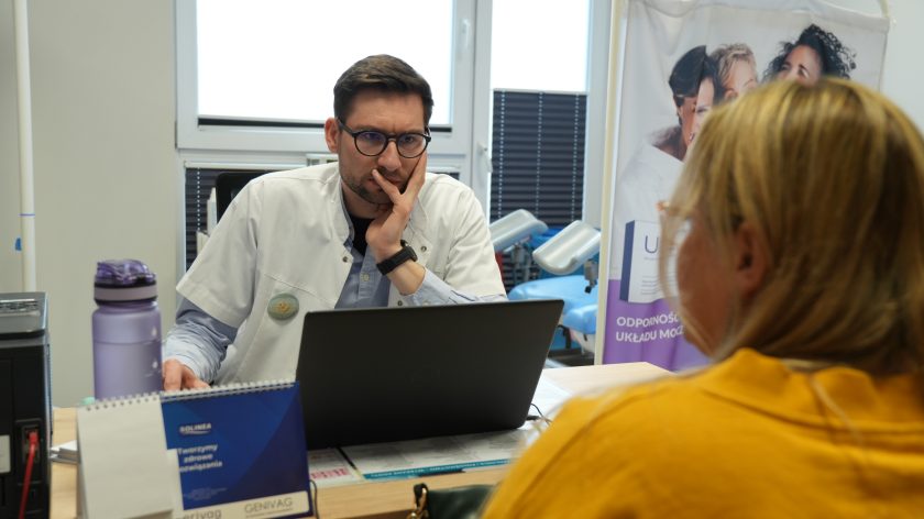 Lekarz w białym fartuchu prowadzi konsultację przy biurku, analizując dane na laptopie, naprzeciwko siedzi pacjentka w żółtej bluzie