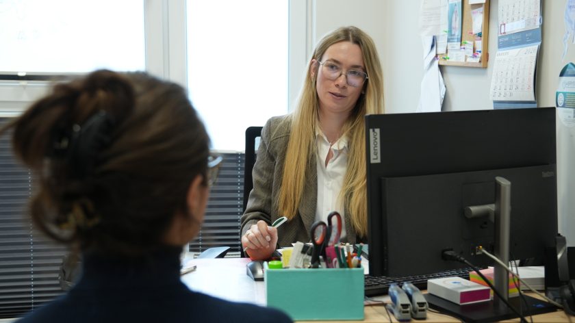 Pracowniczka administracyjna w gabinecie medycznym rozmawia z pacjentką siedzącą naprzeciwko, na biurku komputer i przybory biurowe