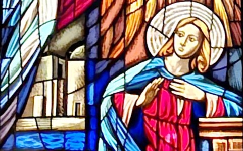 Kolorowy witraż przedstawiający scenę Zwiastowania Najświętszej Maryi Pannie – Maryja w czerwono-niebieskich szatach z aureolą na tle architektury, ukazana w chwili przyjęcia Bożego posłania