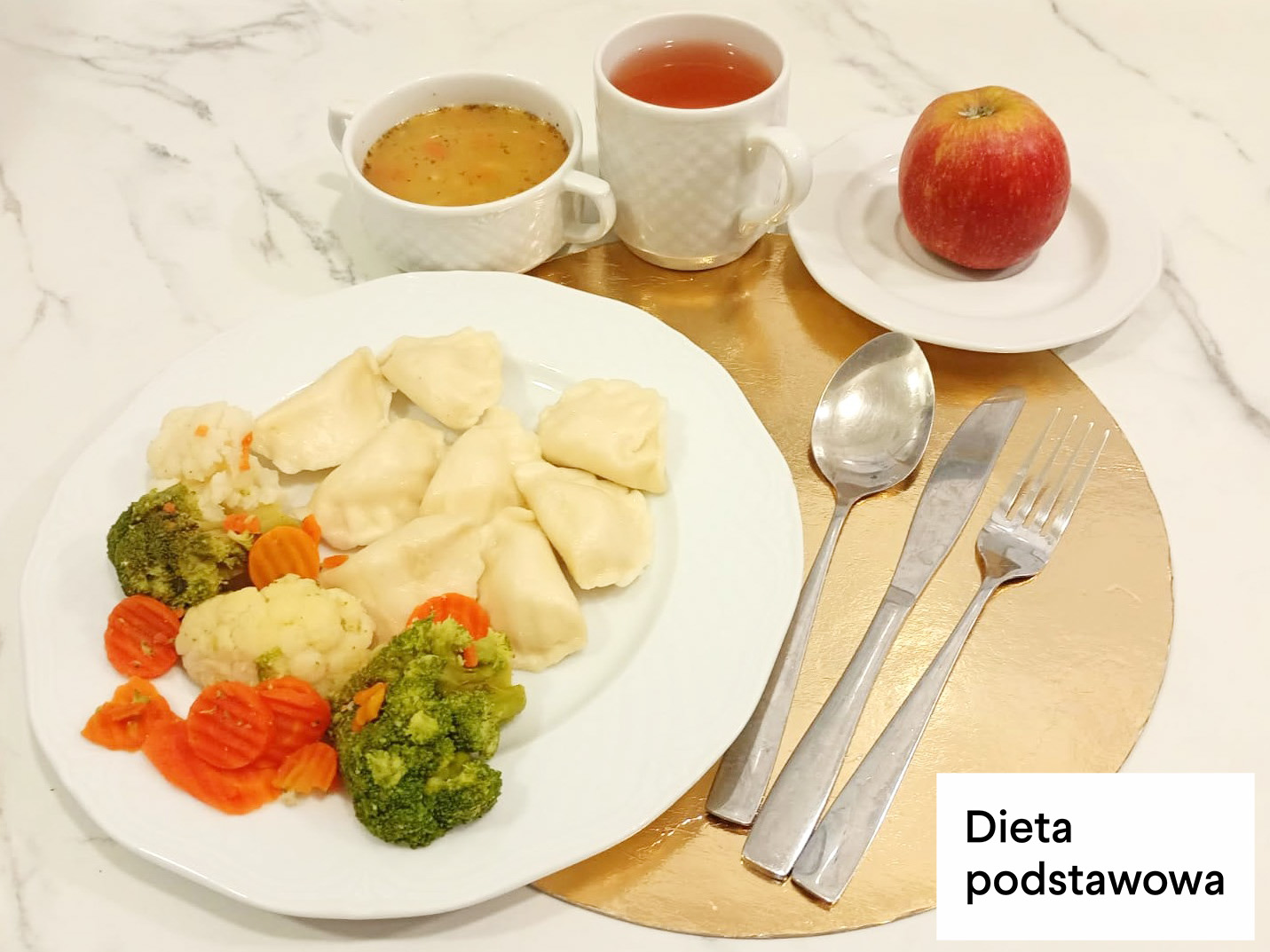 Obiad w diecie podstawowej. Na białym talerzu podano pierogi z farszem oraz porcję gotowanych warzyw – brokułów, kalafiora i marchewki w plasterkach. Obok talerza leżą sztućce. W tle znajdują się filiżanka z lekką zupą oraz kubek z herbatą, a na osobnym talerzyku czerwone jabłko