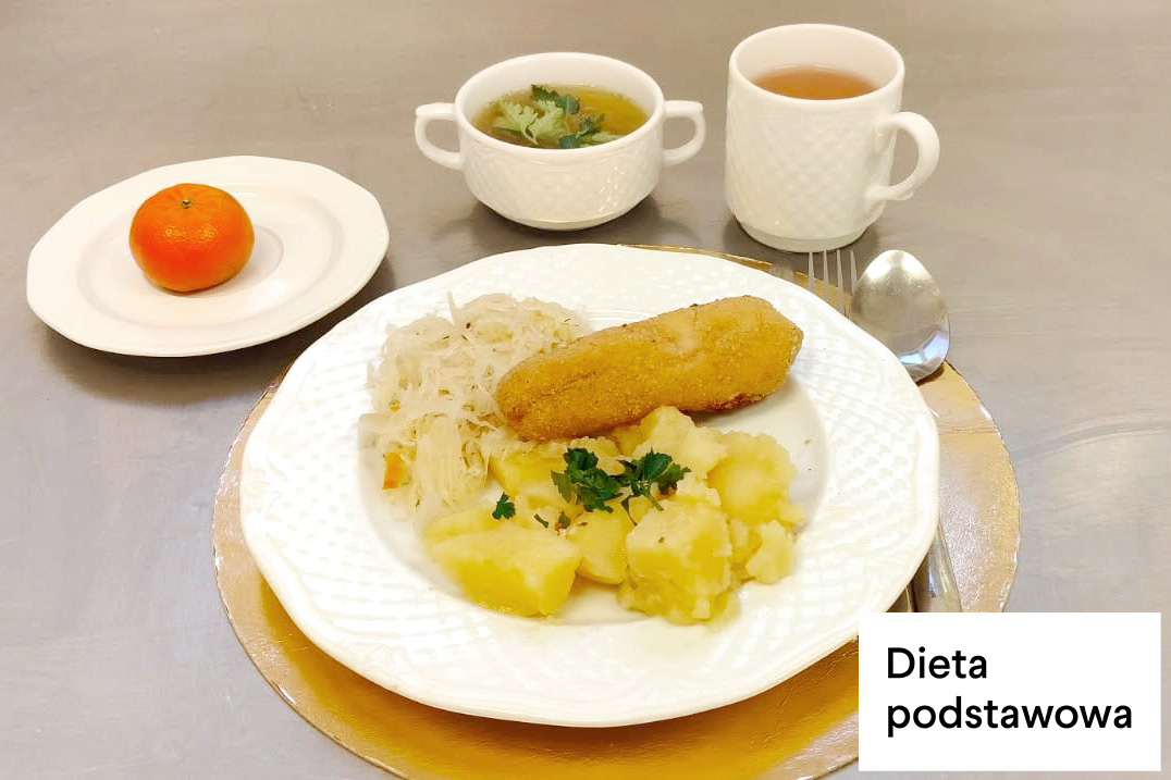 Obiad w diecie podstawowej. Na talerzu podano panierowany filet rybny, gotowane ziemniaki z masłem oraz surówkę z kiszonej kapusty. Obok talerza leży łyżka. W tle stoją filiżanka z zupą jarzynową, kubek z herbatą oraz talerzyk z mandarynką