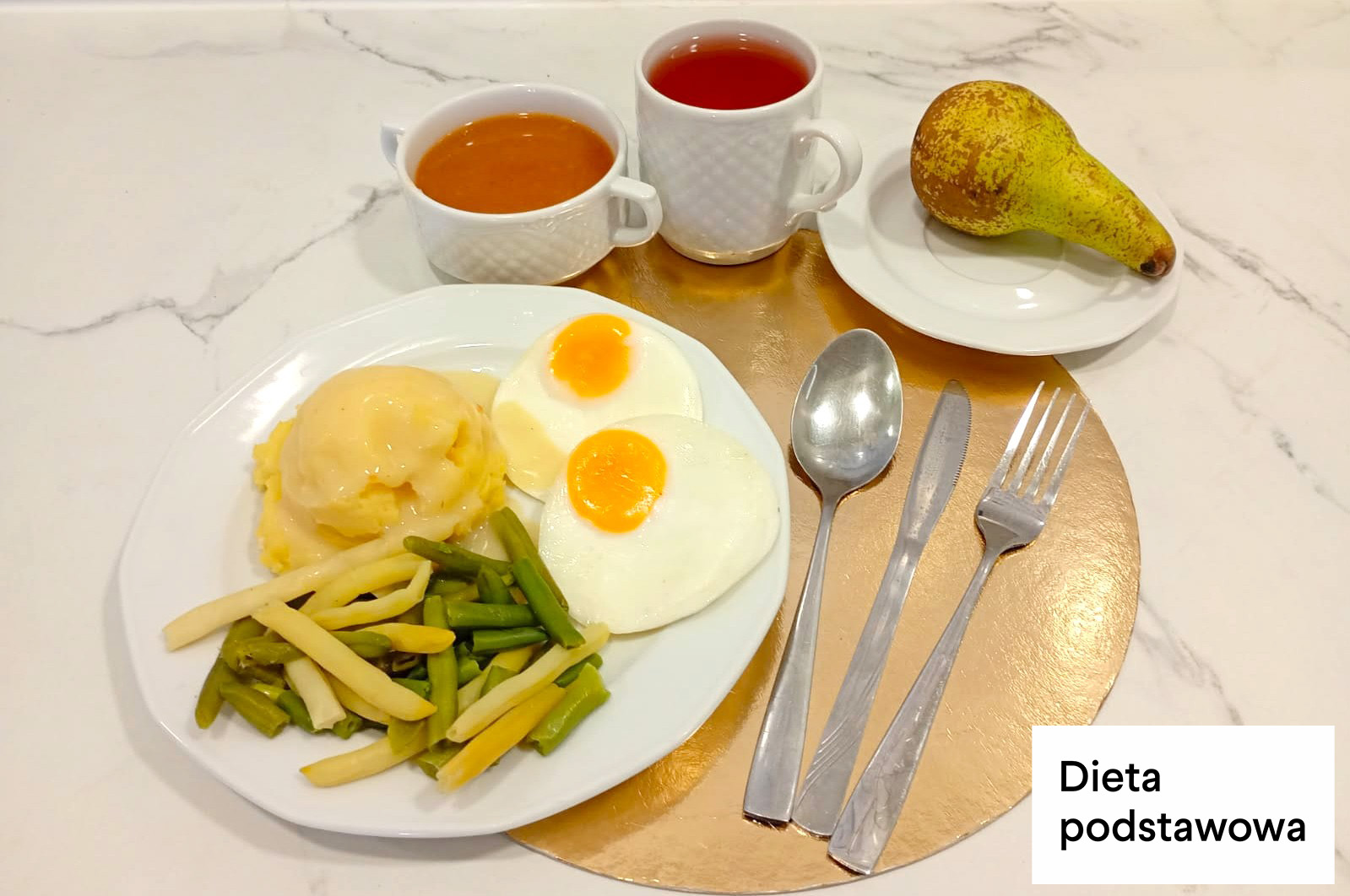 Obiad w diecie podstawowej. Na talerzu podano puree ziemniaczane, dwa jajka sadzone oraz porcję gotowanej fasolki szparagowej. Obok talerza leżą sztućce. W tle znajdują się filiżanka z zupą, kubek z herbatą oraz talerzyk z gruszką