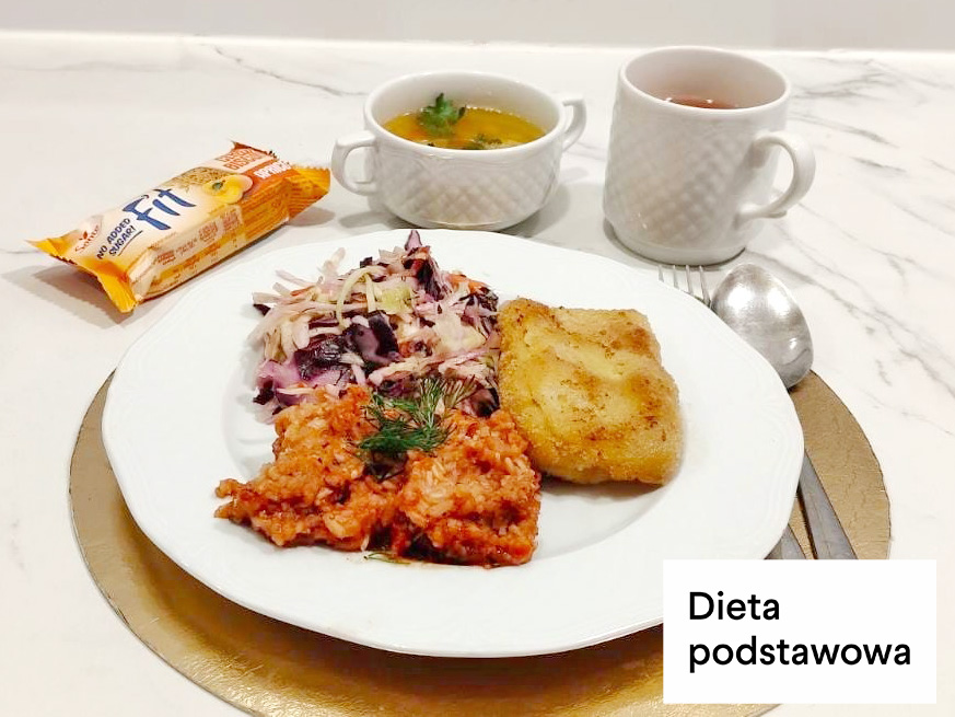 Obiad w diecie podstawowej. Na talerzu znajduje się panierowany filet rybny, porcja ryżu z sosem pomidorowym oraz surówka z kapusty z dodatkiem czerwonej fasoli. W tle ustawiono filiżankę z zupą, kubek z herbatą oraz zapakowane herbatniki