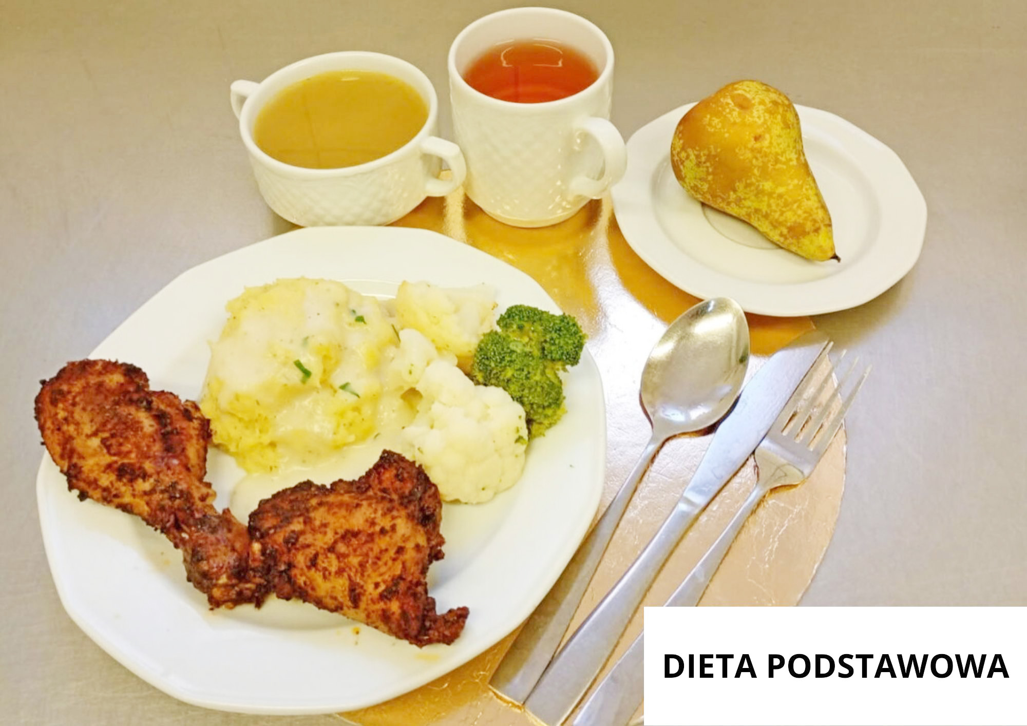 Na białym talerzu podano dwa pieczone kawałki kurczaka, porcję puree ziemniaczanego oraz gotowane warzywa: kalafior i brokuł. Za talerzem stoją dwie białe filiżanki z napojami, a na osobnym talerzyku leży gruszka. Obok nakrycia leżą łyżka, nóż i widelec. W prawym dolnym rogu znajduje się napis „DIETA PODSTAWOWA”