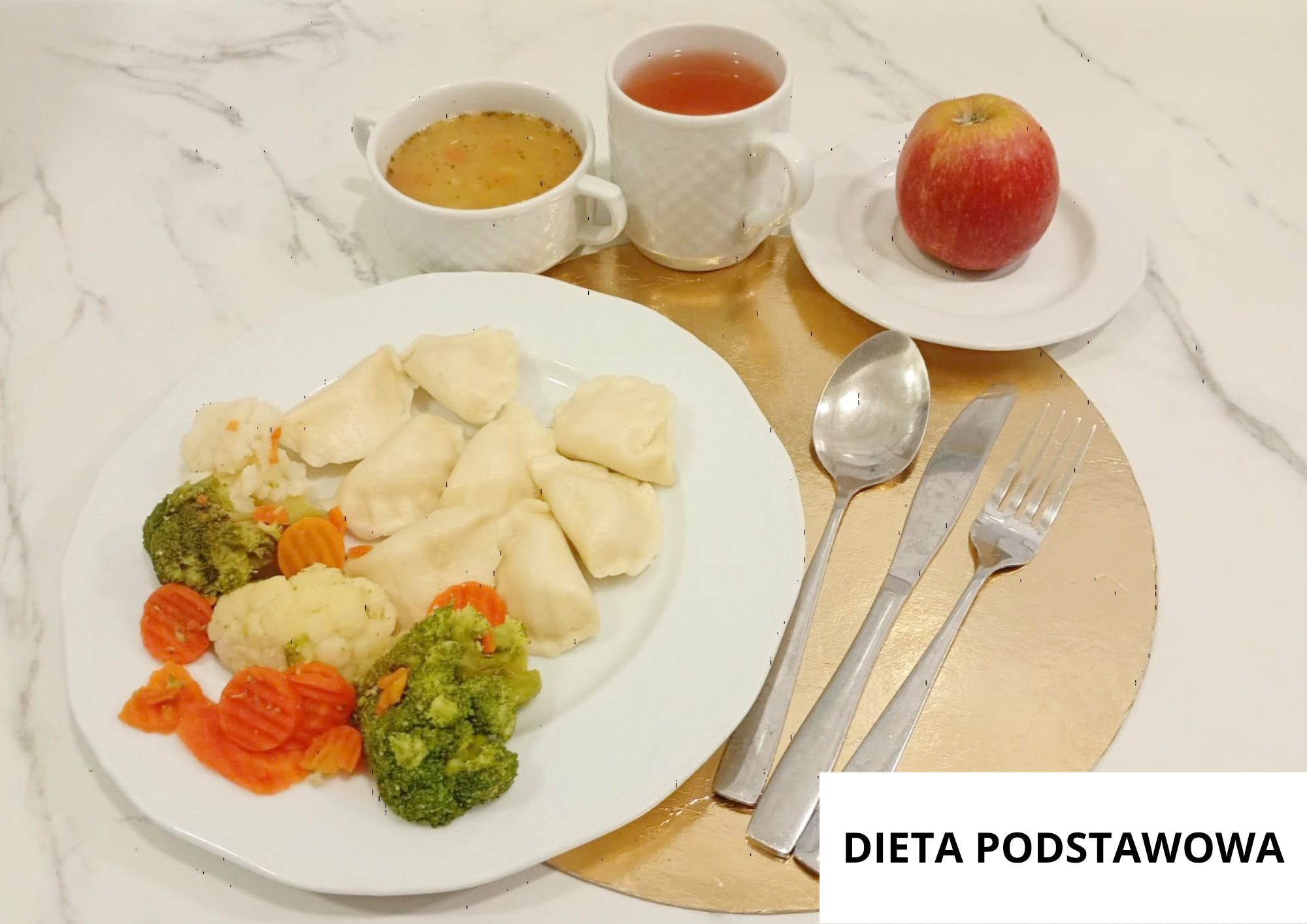 Na białym talerzu znajduje się porcja pierogów oraz gotowane warzywa: marchew, kalafior i brokuły. Nad talerzem stoją dwie białe filiżanki – jedna z zupą, druga z herbatą – a na osobnym talerzyku leży czerwone jabłko. Po prawej stronie leżą łyżka, nóż i widelec. W prawym dolnym rogu widoczny jest napis „DIETA PODSTAWOWA”