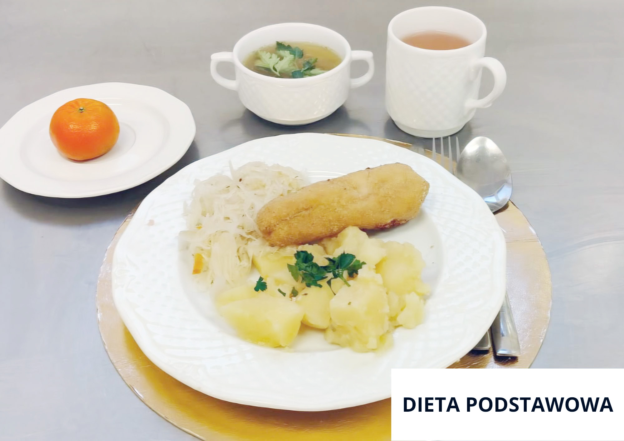 Na białym talerzu podano kotlet panierowany, gotowane ziemniaki i surówkę z kiszonej kapusty. Nad talerzem stoją dwie białe filiżanki – jedna z zupą, druga z herbatą – a po lewej stronie na osobnym talerzyku leży mandarynka. Przy talerzu leżą łyżka i widelec. W prawym dolnym rogu znajduje się napis „DIETA PODSTAWOWA”