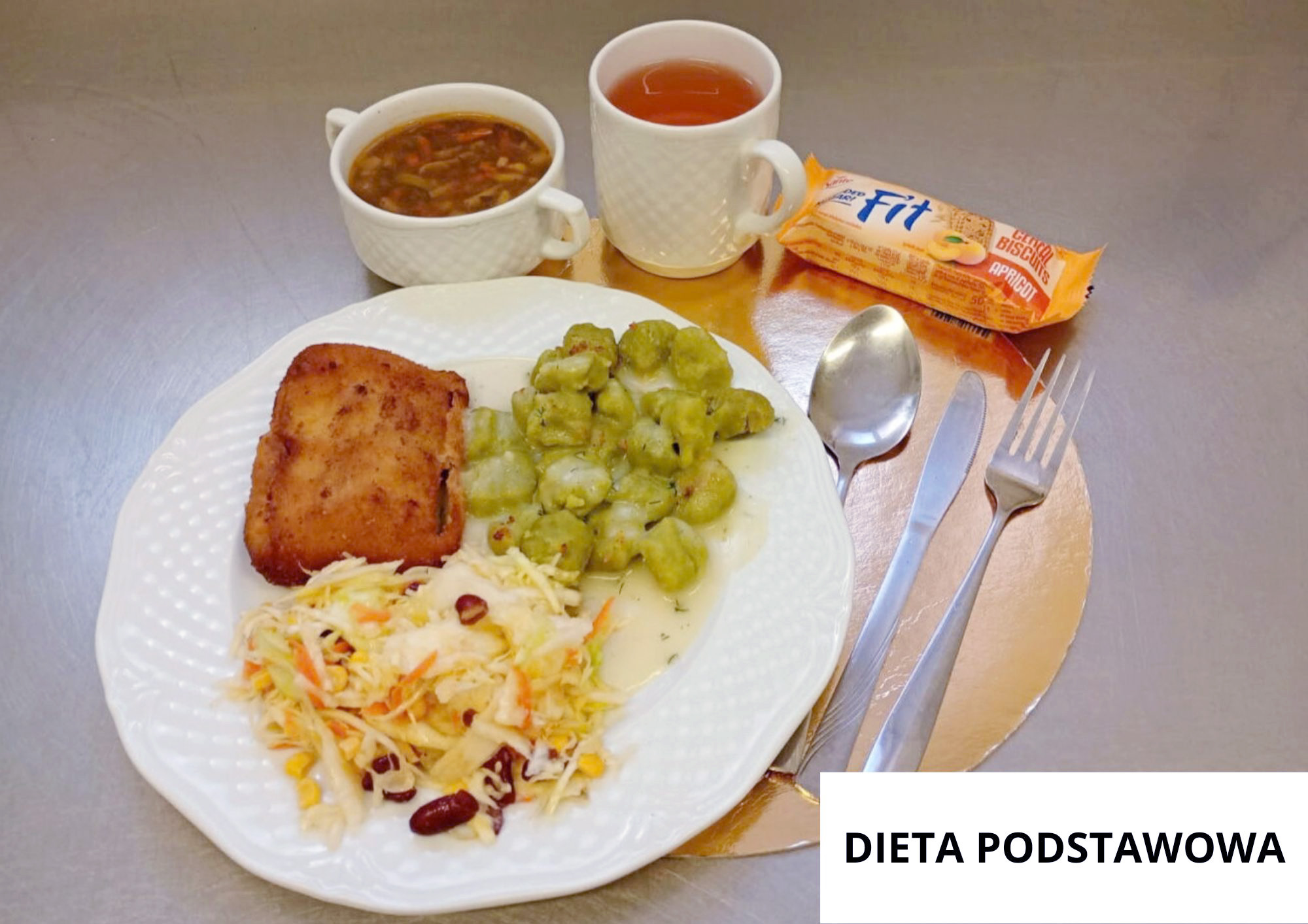 Na białym talerzu leży prostokątny panierowany kotlet, kluski w jasnym sosie oraz surówka z białej kapusty z dodatkiem marchewki, kukurydzy i czerwonej fasoli. Za talerzem stoją dwie białe filiżanki – jedna z zupą, druga z herbatą – oraz opakowanie herbatników. Obok talerza leżą łyżka, nóż i widelec. W prawym dolnym rogu widoczny jest napis „DIETA PODSTAWOWA”