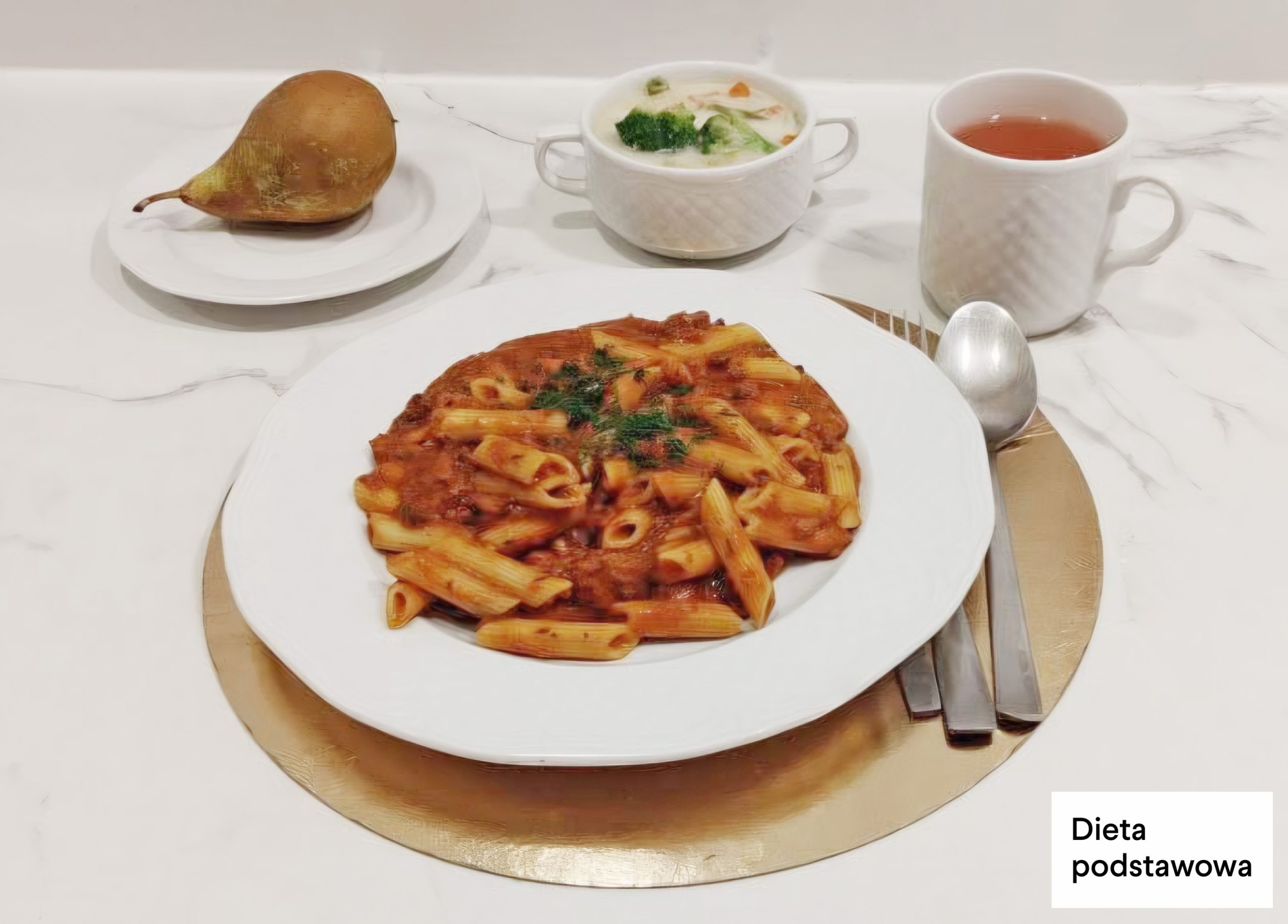 Na białym talerzu ustawionym na złotej podkładce znajduje się porcja makaronu penne w czerwonym sosie pomidorowym, posypana zieloną natką pietruszki. Obok talerza leżą sztućce – nóż i łyżka. W tle, na jasnym blacie, ustawiono białą filiżankę z zupą jarzynową z widocznymi kawałkami brokułu i marchewki oraz biały kubek z herbatą. Po lewej stronie, na małym białym talerzyku, znajduje się świeża gruszka. W prawym dolnym rogu widoczny napis „Dieta podstawowa”