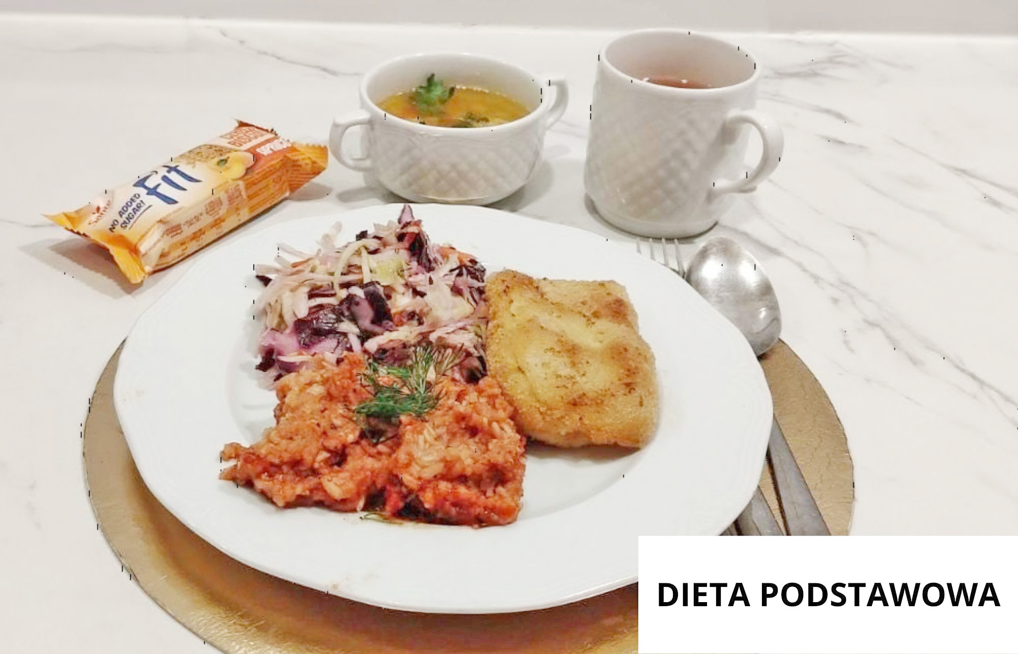 Na białym talerzu znajduje się panierowany kotlet, porcja ryżu w czerwonym sosie oraz surówka z buraczków i kapusty. Nad talerzem stoją dwie białe filiżanki – jedna z zupą, druga z herbatą – a po lewej stronie leży opakowanie herbatników. Obok talerza znajdują się łyżka, nóż i widelec. W prawym dolnym rogu widoczny jest napis „DIETA PODSTAWOWA”