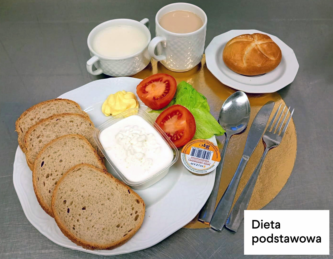 Śniadanie w diecie podstawowej. Na talerzu podano trzy kromki chleba pszenno-żytniego, porcję serka wiejskiego w pojemniku, masło, pomidora oraz liście sałaty. Obok stoją filiżanka z mlekiem i kubek z kawą z mlekiem. Na osobnym talerzyku znajduje się bułka pszenna