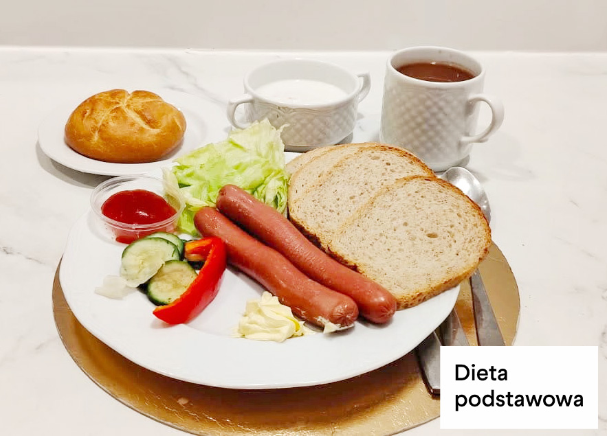Śniadanie w diecie podstawowej. Na talerzu znajdują się dwie parówki drobiowe, kromki chleba pszenno-żytniego, liście sałaty, ogórek, papryka oraz porcja masła. Obok ustawiono mały pojemnik z ketchupem. W tle znajdują się filiżanka z mlekiem, kubek z herbatą oraz bułka pszenna na talerzyku