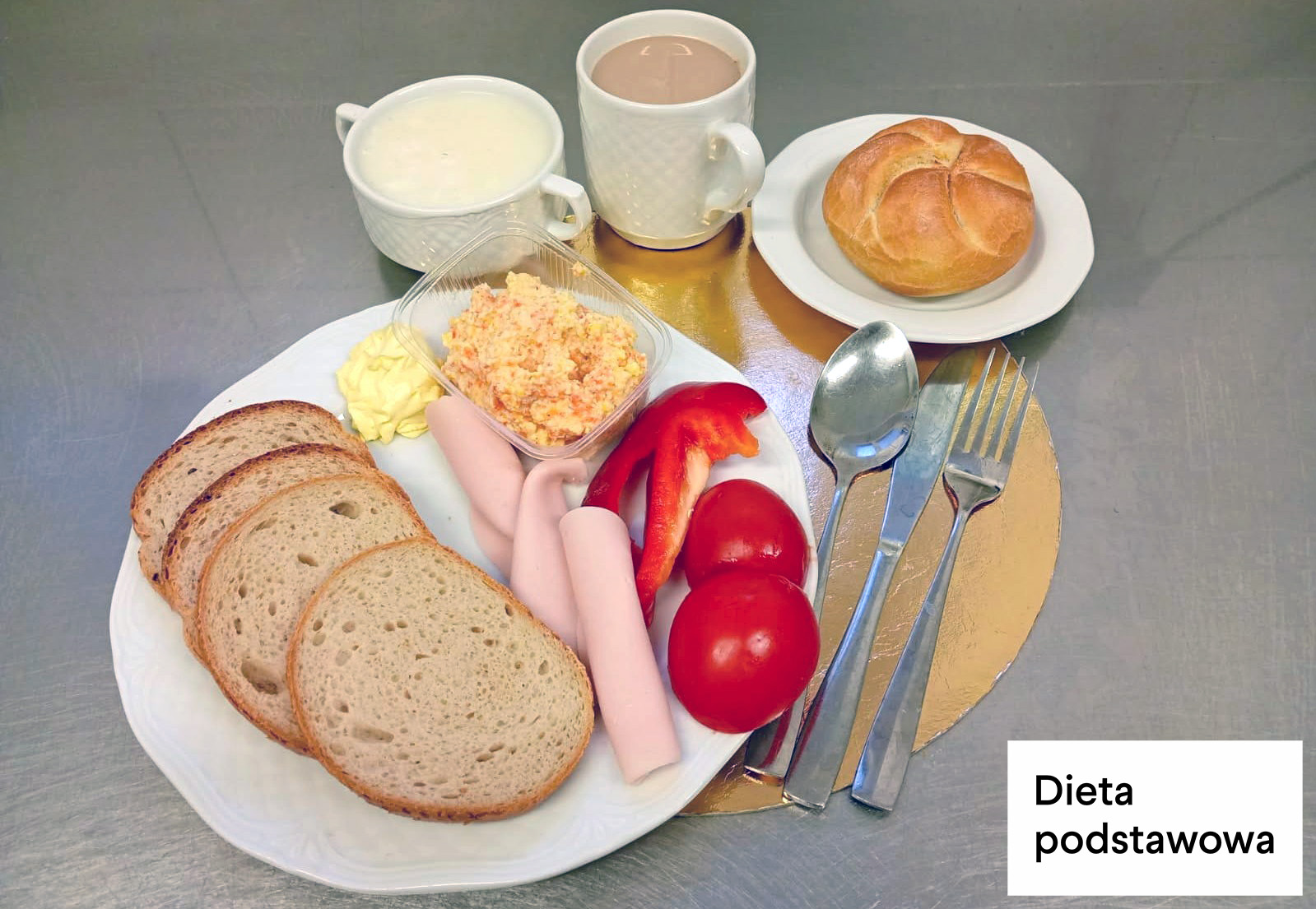 Na stalowym blacie ustawiono zestaw śniadaniowy diety podstawowej. Na dużym białym talerzu znajdują się trzy kromki chleba pszenno-żytniego, dwa plasterki jasnej wędliny drobiowej, porcja sałatki jarzynowej w przezroczystym pojemniku, porcja masła, kawałki czerwonej papryki oraz dwa pomidory. Obok talerza leżą sztućce. Na małych talerzykach ustawiono bułkę pszenną, kubek z kawą z mlekiem oraz filiżankę z mlekiem