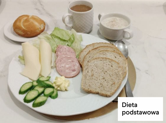 Śniadanie w diecie podstawowej. Na talerzu podano trzy kromki chleba pszenno-żytniego, plaster sera żółtego, plaster sera typu mozzarella, kilka plastrów wędliny oraz świeże warzywa – ogórek i liście sałaty. Na talerzu znajduje się także porcja masła. W tle stoją kubek z kawą z mlekiem, filiżanka z mlekiem oraz talerzyk z bułką pszenną