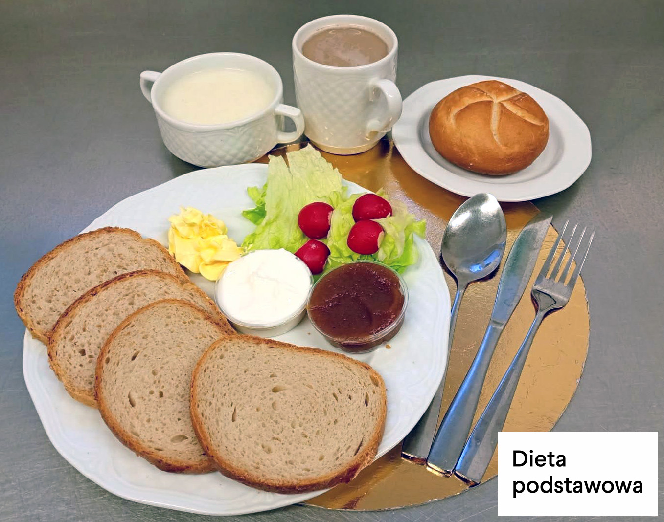 Śniadanie w diecie podstawowej. Na talerzu znajdują się trzy kromki chleba pszenno-żytniego, porcja twarogu w pojemniku, porcja dżemu w małym pojemniku, masło oraz świeże warzywa – liście sałaty i rzodkiewki. Obok talerza leżą sztućce. W tle stoją filiżanka z mlekiem, kubek z kawą z mlekiem oraz bułka pszenna na talerzyku