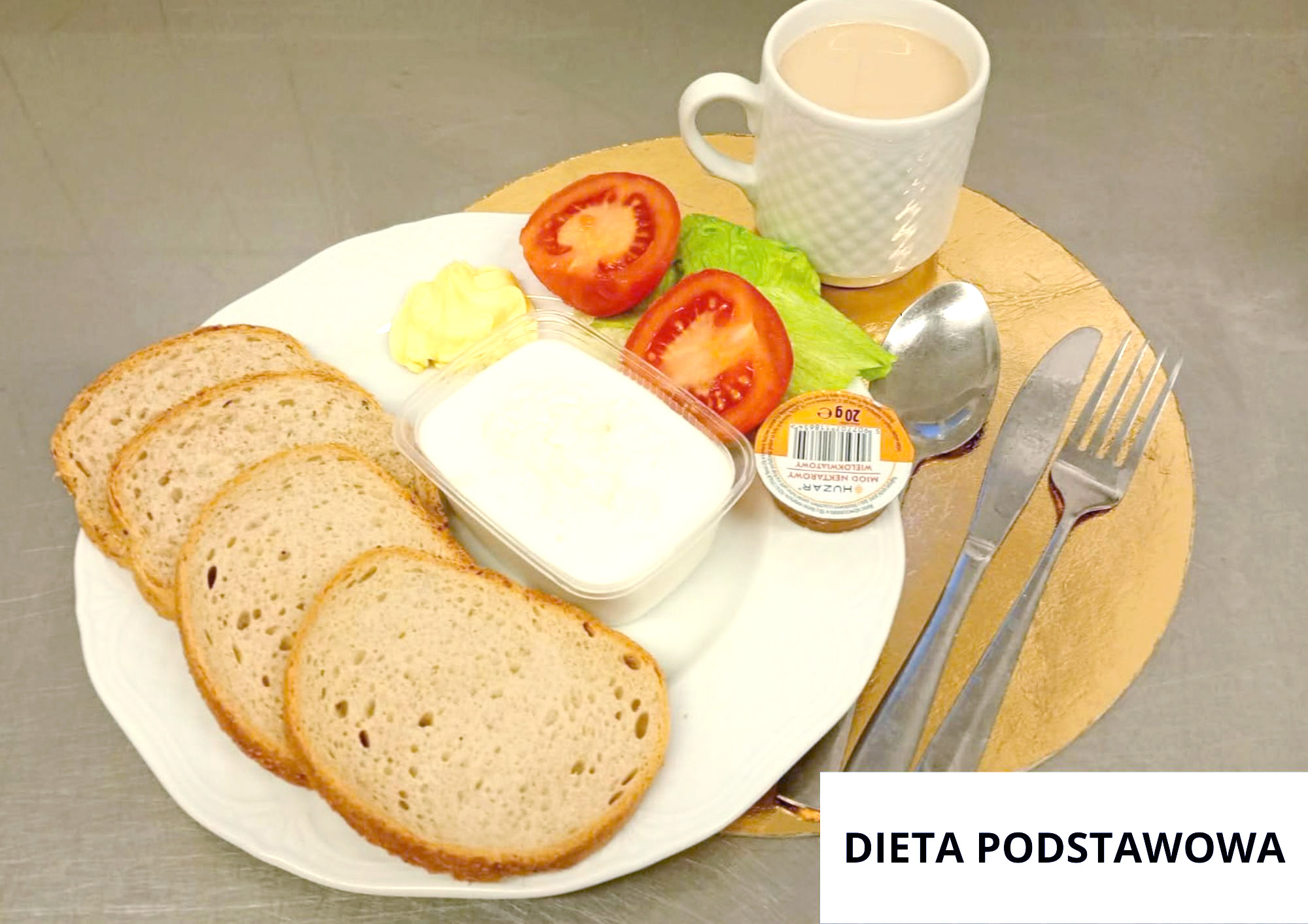 Na białym talerzu leżą cztery kromki chleba, pojemnik z białym twarożkiem, mały zamknięty pojemniczek, dwa kawałki pomidora, liść sałaty i porcja masła. Za talerzem stoi biały kubek z jasnym napojem. Po prawej stronie leżą łyżka, nóż i widelec. W prawym dolnym rogu znajduje się napis „DIETA PODSTAWOWA”