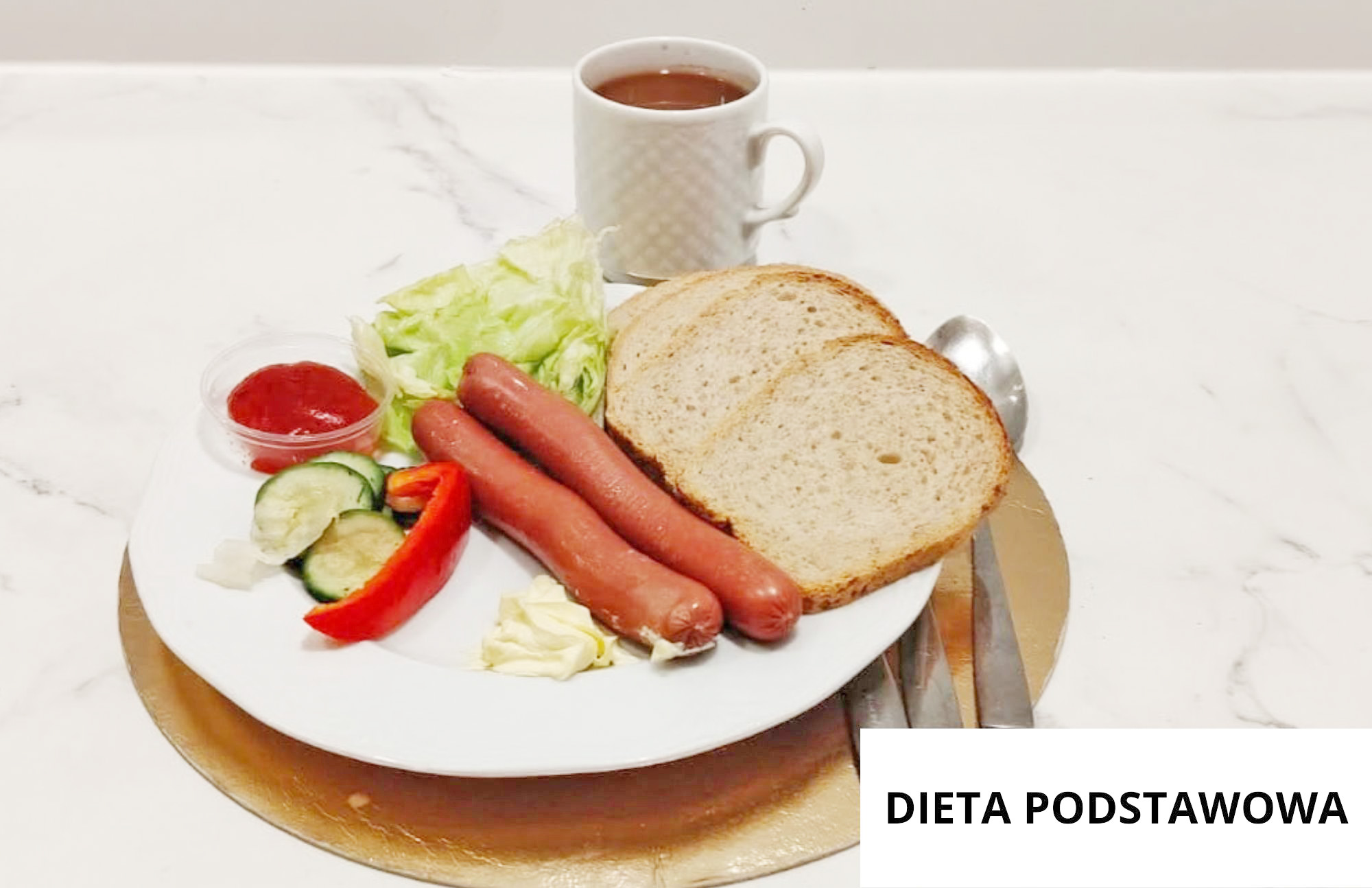 Na białym talerzu podano dwie parówki, dwie kromki chleba, liście sałaty, plasterki ogórka, kawałek czerwonej papryki, porcję masła oraz mały pojemnik z ketchupem. Za talerzem stoi biały kubek z herbatą. Obok leżą łyżka i nóż. W prawym dolnym rogu widoczny jest napis „DIETA PODSTAWOWA”