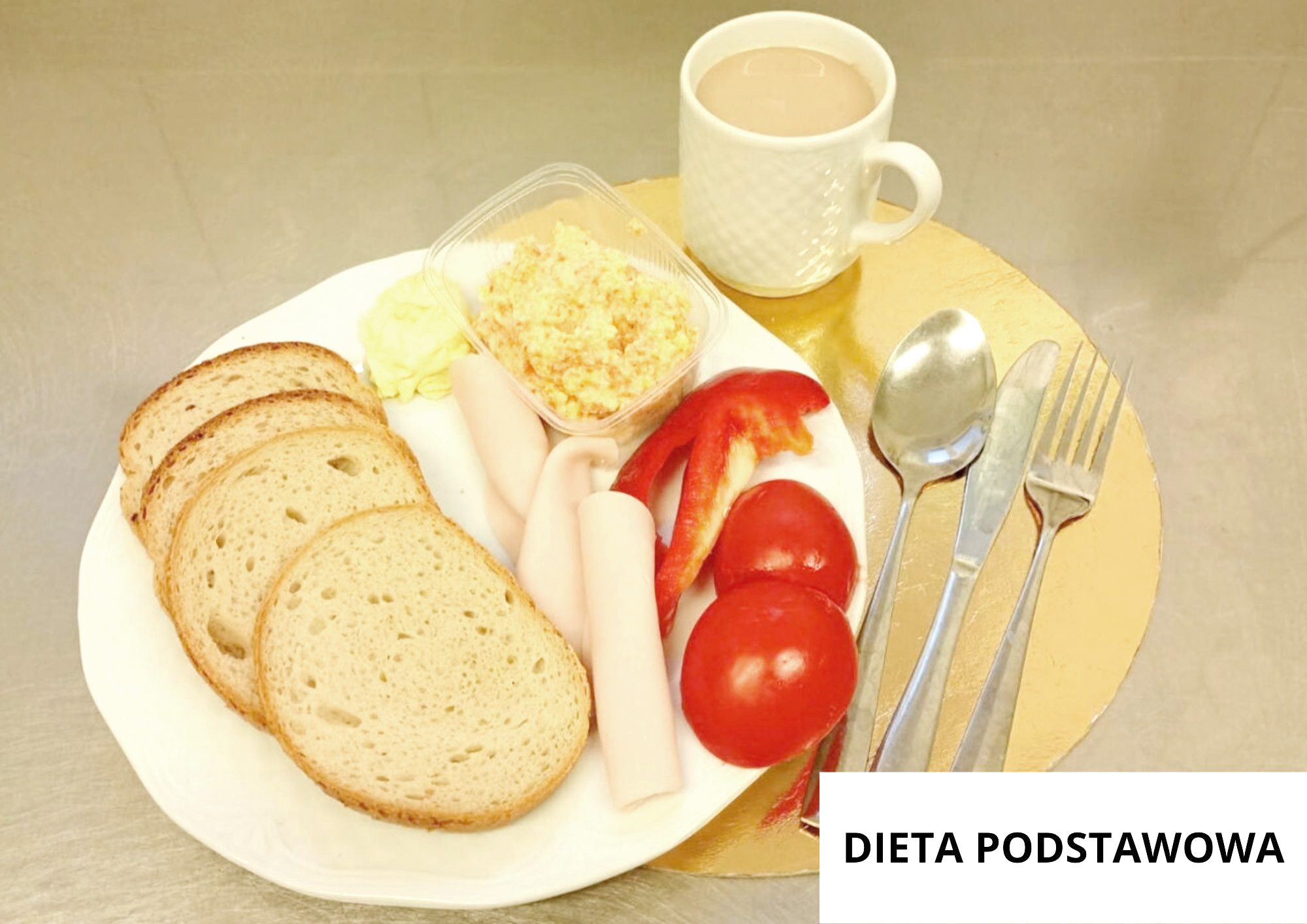 Na białym talerzu leżą cztery kromki chleba, dwa jasne plasterki wędliny zrolowane w rulon, pojemnik z pastą jajeczną, dwa pomidory, kawałek czerwonej papryki oraz porcja masła. Za talerzem stoi biały kubek z jasnym napojem. Po prawej stronie leżą łyżka, nóż i widelec. W prawym dolnym rogu znajduje się napis „DIETA PODSTAWOWA”