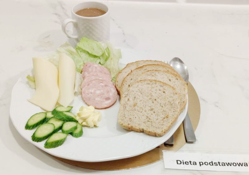 Na białym talerzu leżą trzy kromki chleba, dwa plastry jasnego sera, trzy plastry wędliny, liście sałaty, kilka plasterków ogórka i porcja masła. Obok talerza stoi biały kubek z jasnym napojem. Przy talerzu leżą łyżka i nóż, a w prawym dolnym rogu widoczny jest napis „Dieta podstawowa”