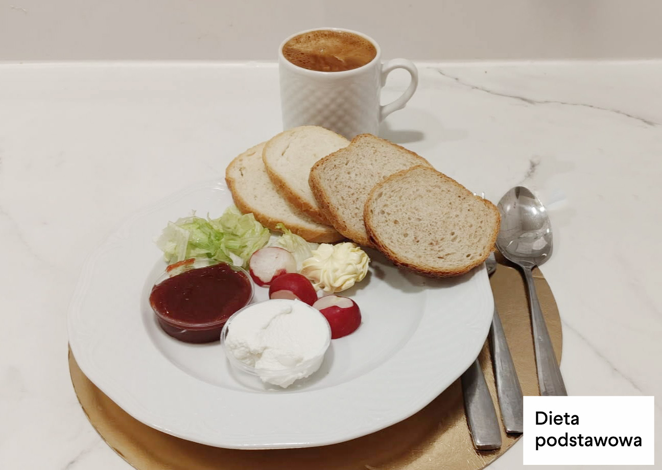 Na białym talerzu umieszczonym na złotej podkładce znajdują się cztery kromki jasnego pieczywa. Obok pieczywa podano porcję białego twarogu, rozetkę masła oraz niewielką porcję sałaty i plasterki rzodkiewki. Na talerzu znajduje się także mały pojemnik z dżemem. Za talerzem ustawiono biały kubek z kawą. Po prawej stronie leżą łyżka i widelec. W prawym dolnym rogu widnieje napis „Dieta podstawowa”