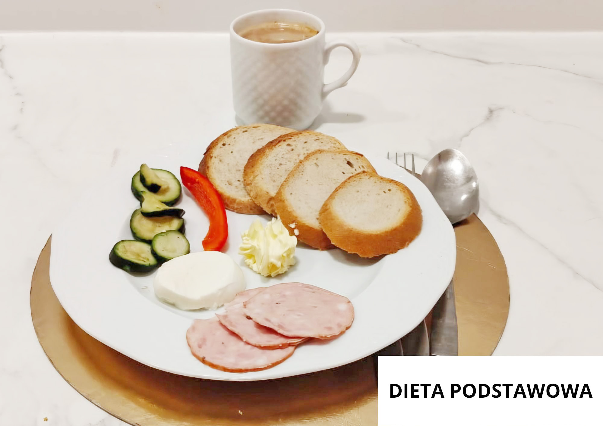 Na białym talerzu położonym na złotej podkładce znajdują się cztery kromki jasnego pieczywa. Obok pieczywa ułożono plasterki szynki, porcję białego sera, rozetkę masła oraz świeże warzywa: plasterki ogórka i kawałek czerwonej papryki. Za talerzem stoi biały kubek z kawą. Po prawej stronie leżą łyżka i widelec. W prawym dolnym rogu widnieje napis „DIETA PODSTAWOWA”