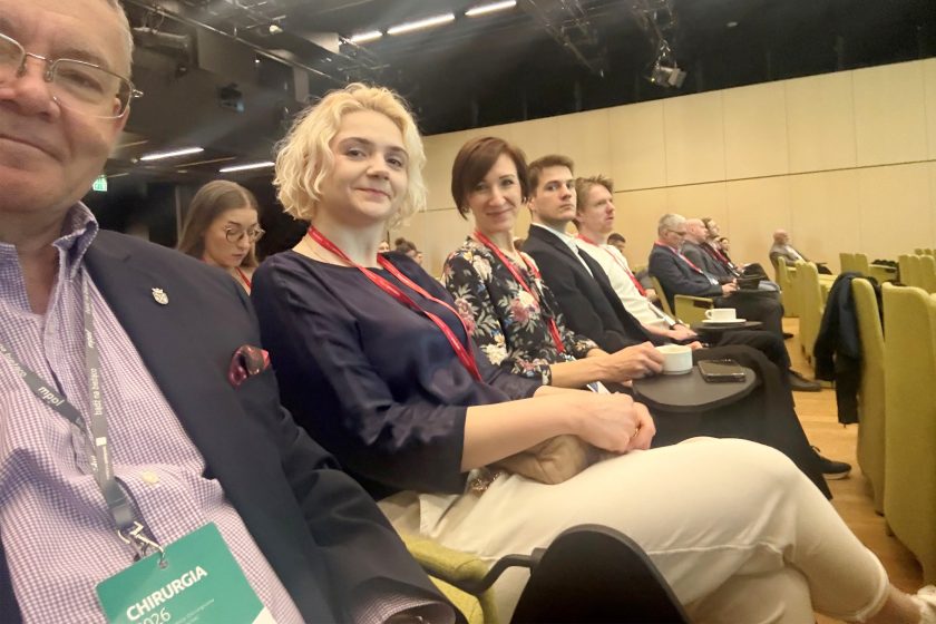 Uczestnicy konferencji siedzą na widowni sali wykładowej. Na pierwszym planie kilka osób z identyfikatorami konferencyjnymi, w tym kobiety i mężczyźni, obserwują wydarzenie; część z nich spogląda w stronę aparatu. W tle widoczne rzędy siedzeń oraz inni uczestnicy