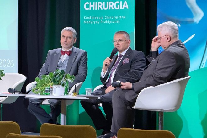 Panel dyskusyjny podczas Konferencji „Chirurgia 2026”. Trzech mężczyzn w garniturach siedzi na scenie w fotelach, jeden z nich trzyma mikrofon i zabiera głos. W tle zielona plansza z nazwą konferencji, przed nimi stolik z wodą i rośliną