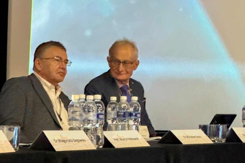 Dwóch uczestników konferencji medycznej siedzi przy stole prezydialnym podczas panelu dyskusyjnego. Mężczyźni ubrani są w garnitury i identyfikatory konferencyjne. Na stole przed nimi stoją butelki wody mineralnej, szklanki oraz tabliczki z nazwiskami prelegentów. W tle widoczny jest ekran projekcyjny z rozmytym obrazem prezentacji