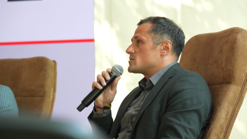 Mężczyzna w ciemnym garniturze siedzi w fotelu panelowym i wypowiada się do mikrofonu podczas konferencji. W tle widoczny jest duży ekran prezentacyjny z grafikami związanymi z Bonifraterskim Centrum Medycznym. Uczestnik panelu zwrócony jest w stronę innych prelegentów i publiczności