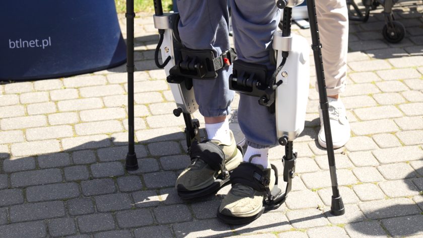 Detal urządzenia rehabilitacyjnego typu egzoszkielet zamocowanego na nogach uczestnika pokazu. Widoczne są elementy wspomagające ruch kończyn dolnych oraz podpórki pomagające w chodzeniu podczas rehabilitacji