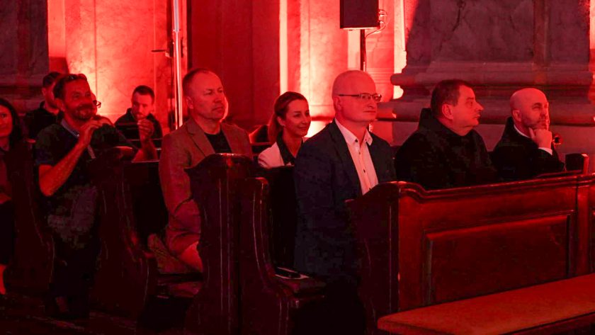 Uczestnicy koncertu siedzą w ławkach zabytkowego kościoła, wsłuchując się w występ artystów. Wnętrze świątyni rozświetla czerwona iluminacja podkreślająca architekturę i wyjątkowy charakter wydarzenia