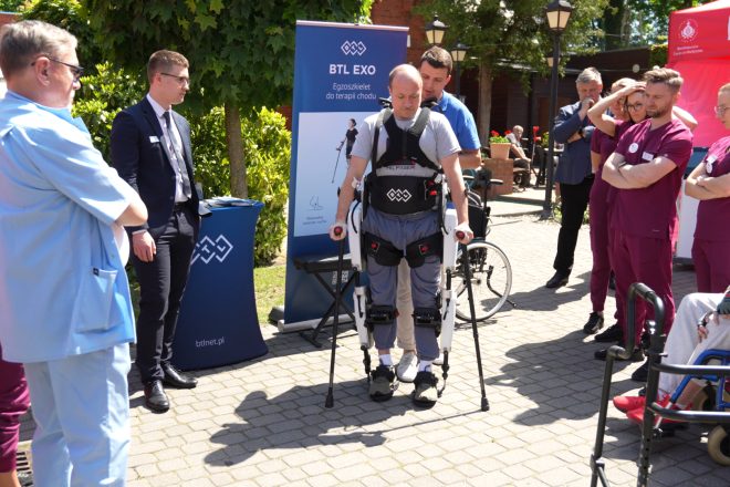 Pokaz działania egzoszkieletu rehabilitacyjnego podczas wydarzenia plenerowego przy Bonifraterskim Centrum Medycznym. Uczestnik wyposażony w urządzenie wspomagające chód porusza się przy wsparciu instruktora, a wokół zgromadzili się pracownicy medyczni i obserwatorzy