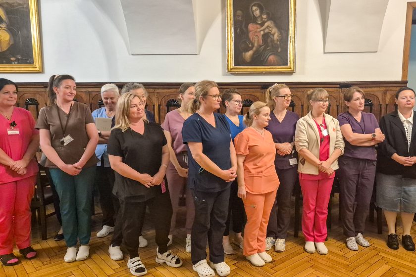 Grupa pielęgniarek i pracownic medycznych stoi w zabytkowej sali szpitala Bonifratrów w Krakowie. Kobiety ubrane są w kolorowe stroje medyczne – granatowe, różowe, fioletowe, pomarańczowe i zielone. W tle widoczne są drewniane ściany oraz obrazy religijne w złotych ramach, podkreślające historyczny charakter wnętrza