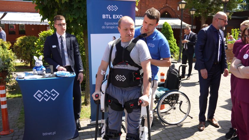 Mężczyzna korzystający z egzoszkieletu rehabilitacyjnego stoi na dziedzińcu podczas prezentacji technologii wspomagającej terapię chodu. Obok znajduje się instruktor oraz stoisko firmy prezentującej sprzęt rehabilitacyjny