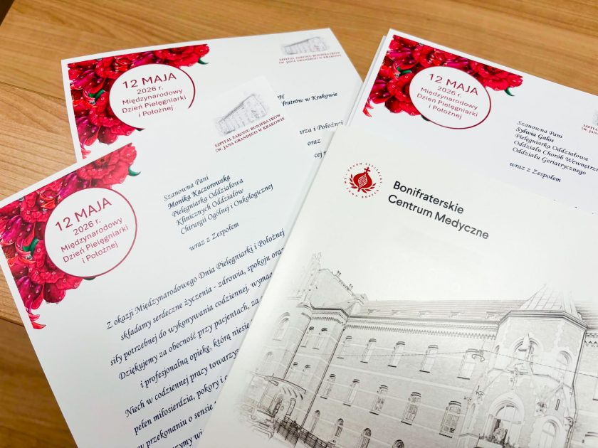 Na drewnianym stole rozłożone są okolicznościowe dyplomy przygotowane z okazji Międzynarodowego Dnia Pielęgniarki i Położnej oraz publikacja Bonifraterskiego Centrum Medycznego przedstawiająca Szpital Zakonu Bonifratrów św. Jana Grandego w Krakowie. Dokumenty ozdobione są czerwonym motywem kwiatowym i zawierają indywidualne podziękowania dla personelu medycznego