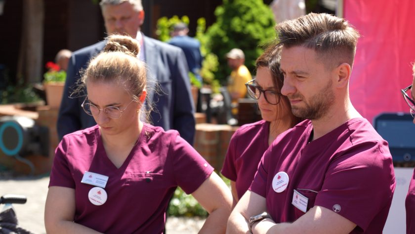 Grupa pracowników medycznych w bordowych uniformach obserwuje pokaz rehabilitacyjny odbywający się na świeżym powietrzu. Uczestnicy wydarzenia z uwagą śledzą prezentację nowoczesnych rozwiązań wspierających rehabilitację pacjentów