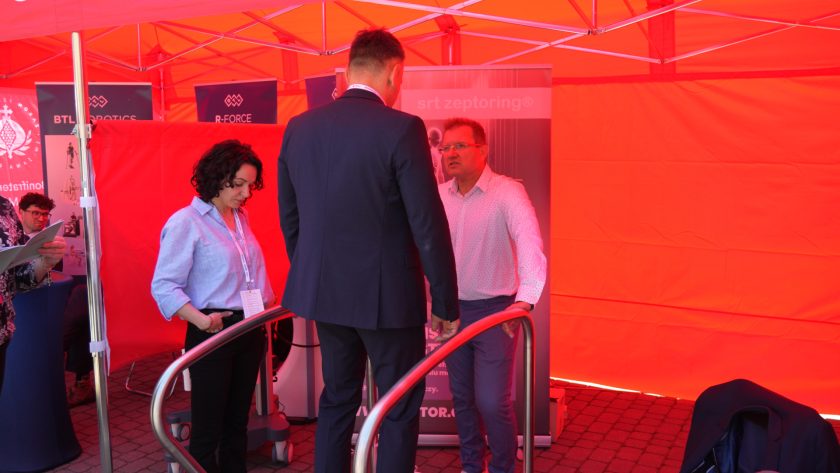 Wnętrze namiotu wystawowego podczas prezentacji urządzeń rehabilitacyjnych. Kilka osób rozmawia przy stoisku prezentującym nowoczesne technologie wspomagające terapię i diagnostykę pacjentów