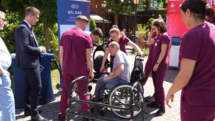 Uczestnik pokazu siedzi na wózku inwalidzkim po zakończeniu demonstracji egzoszkieletu rehabilitacyjnego. Wokół niego znajdują się pracownicy medyczni oraz osoby obserwujące przebieg prezentacji sprzętu wspomagającego rehabilitację ruchową