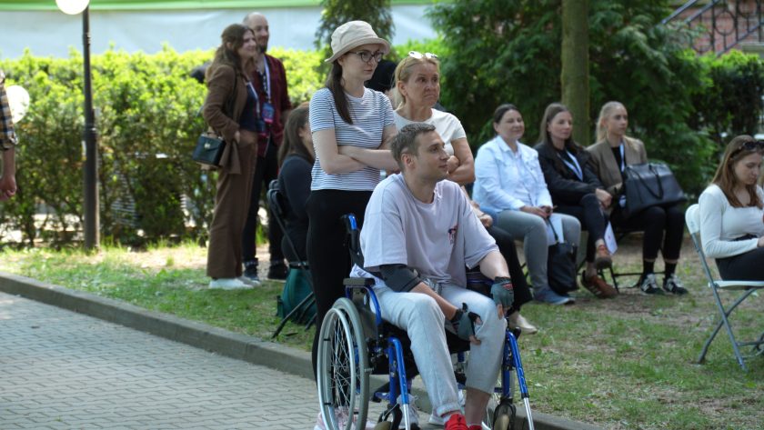 Młody mężczyzna siedzący na wózku inwalidzkim obserwuje wydarzenie odbywające się w plenerze. Obok stoją uczestniczki konferencji i osoby zgromadzone na terenie Bonifraterskiego Centrum Medycznego