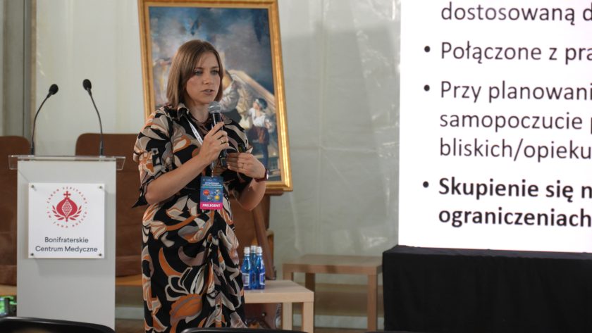 Prelegentka występuje podczas konferencji i przemawia do mikrofonu na tle prezentacji multimedialnej. Obok widoczny jest pulpit z logo Bonifraterskiego Centrum Medycznego oraz slajd dotyczący rehabilitacji i opieki nad pacjentem