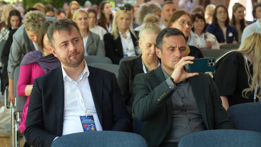 Kobieta w jasnej marynarce prowadzi wykład podczas konferencji organizowanej przez Zakon Szpitalny św. Jana Bożego. Prelegentka przemawia do mikrofonu na tle baneru z logo Zakonu Bonifratrów