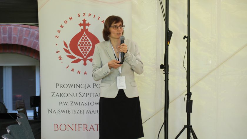 Dwóch uczestników konferencji siedzi wśród publiczności. Jeden z nich wykonuje zdjęcie lub nagranie telefonem komórkowym, a w tle widoczni są pozostali uczestnicy wydarzenia słuchający prelekcji