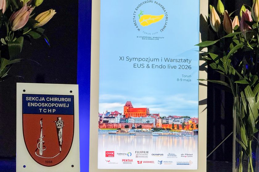Roll-up promujący „XI Sympozjum i Warsztaty EUS & Endo live 2026” ustawiony jest obok emblematu Sekcji Chirurgii Endoskopowej TChP. Na plakacie znajduje się panorama Torunia z podświetloną starówką oraz informacje o wydarzeniu odbywającym się w dniach 8–9 maja 2026 roku. Po bokach kompozycji ustawione są dekoracyjne lilie i zielone rośliny, tworzące elegancką oprawę konferencyjną