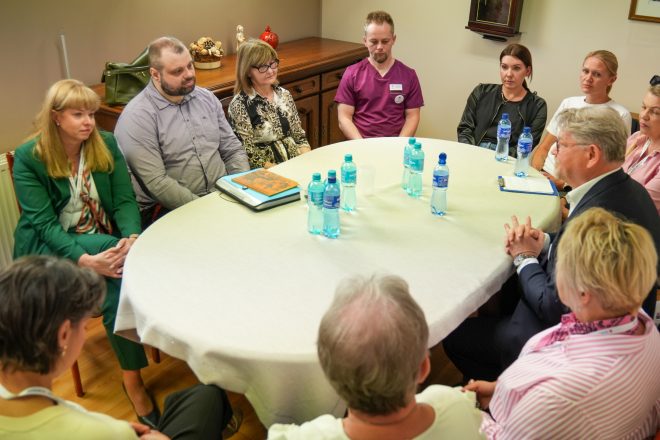 Przy dużym, okrągłym stole nakrytym jasnym obrusem siedzi kilkanaście osób uczestniczących w spotkaniu i dyskusji. W centrum kadru widoczni są przedstawiciele personelu medycznego oraz uczestnicy wydarzenia, siedzący w skupieniu i słuchający wypowiedzi mężczyzny w garniturze znajdującego się po prawej stronie zdjęcia. Na stole stoją butelki wody mineralnej oraz notatnik. W tle znajduje się drewniana komoda i dekoracje wnętrza sali konferencyjnej. Atmosfera spotkania jest spokojna i formalna