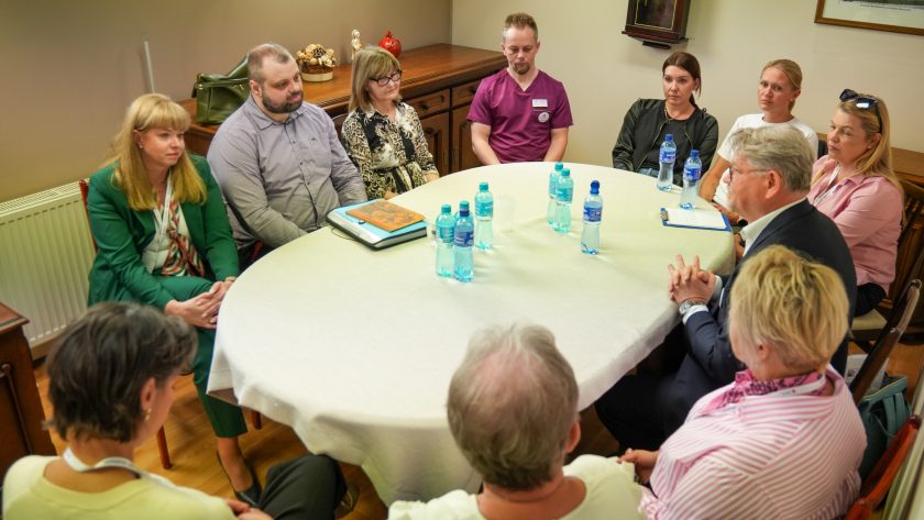 Przy dużym, okrągłym stole nakrytym jasnym obrusem siedzi kilkanaście osób uczestniczących w spotkaniu i dyskusji. W centrum kadru widoczni są przedstawiciele personelu medycznego oraz uczestnicy wydarzenia, siedzący w skupieniu i słuchający wypowiedzi mężczyzny w garniturze znajdującego się po prawej stronie zdjęcia. Na stole stoją butelki wody mineralnej oraz notatnik. W tle znajduje się drewniana komoda i dekoracje wnętrza sali konferencyjnej. Atmosfera spotkania jest spokojna i formalna