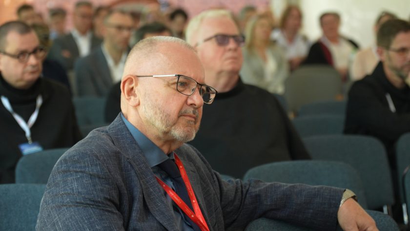 Uczestnik konferencji w okularach i granatowej marynarce siedzi wśród publiczności i słucha wystąpień panelowych. W tle widoczni są inni uczestnicy wydarzenia, w tym duchowni oraz osoby świeckie zgromadzone na sali konferencyjnej
