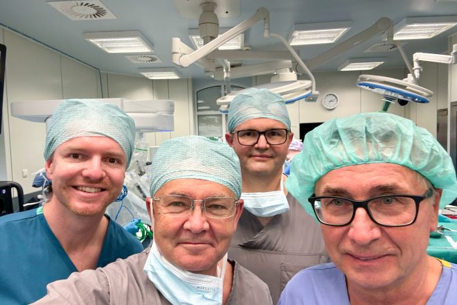 Czterech członków zespołu medycznego pozuje do wspólnego zdjęcia na sali operacyjnej. Lekarze ubrani są w stroje chirurgiczne, czepki i częściowo opuszczone maseczki. W tle widoczne są lampy operacyjne oraz elementy robota chirurgicznego przygotowanego do zabiegu