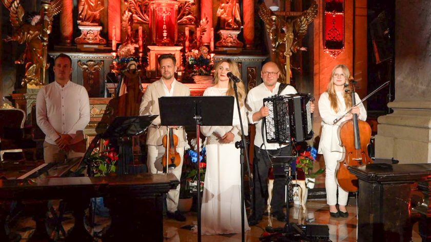 Artyści zespołu muzycznego stoją przed ołtarzem po zakończonym utworze. W skład zespołu wchodzą wokalistka oraz instrumentaliści grający na akordeonie, wiolonczeli, skrzypcach i instrumentach klawiszowych