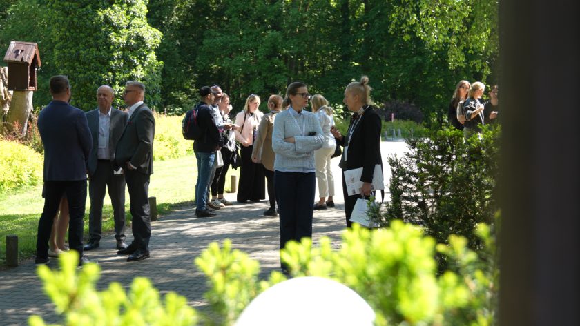 Uczestnicy wydarzenia rozmawiają w plenerze podczas przerwy konferencyjnej. Grupy osób stoją na alejce w otoczeniu zieleni i drzew, prowadząc swobodne rozmowy networkingowe